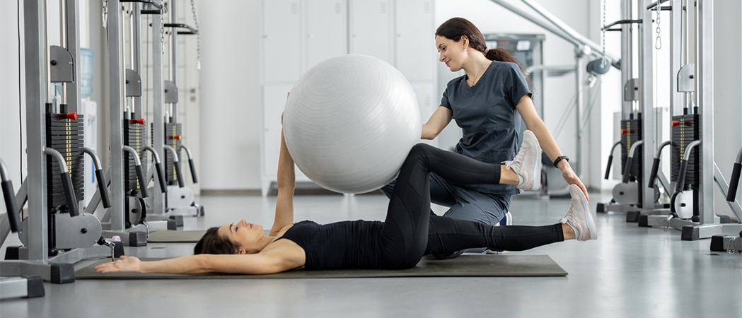 Een vrouw op de grond met een grijze fitnessball met naast haar een vrouwelijke coach
